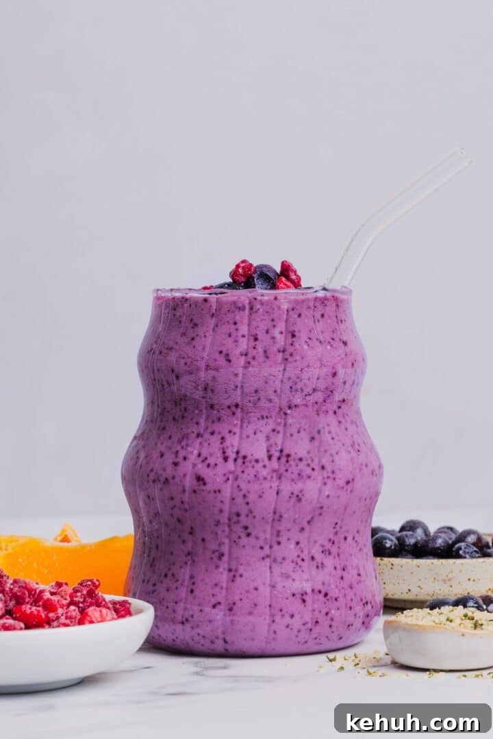Blueberry raspberry smoothie in an etched glass sitting near fruit and other ingredients