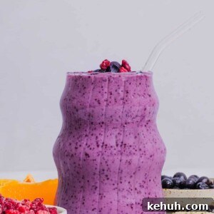 Blueberry raspberry smoothie in an etched glass sitting near fruit and other ingredients