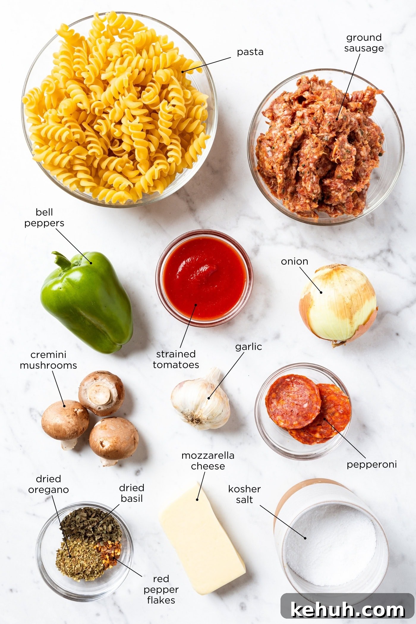 Cheesy Pizza Casserole Recipe with Sausage and Peppers 4 White marble surface with ingredients for pizza casserole including pasta, pepperoni, sauce, veggies, spices, and more.