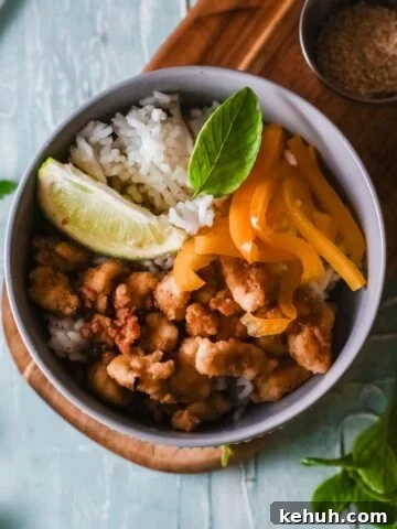 Grey bowl with crispy chicken bites, orange peppers, and rice with lime in the bowl on a wooden cutting board.
