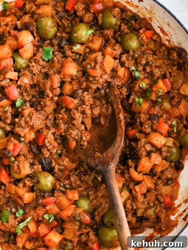 Authentic Cuban Ropa Vieja Recipe — Slow-Cooked Shredded Beef 16 A wooden serving spoon rests inside a simmering pot of Cuban picadillo with ground beef, olives, raisins, and potatoes in a rich tomato sauce.