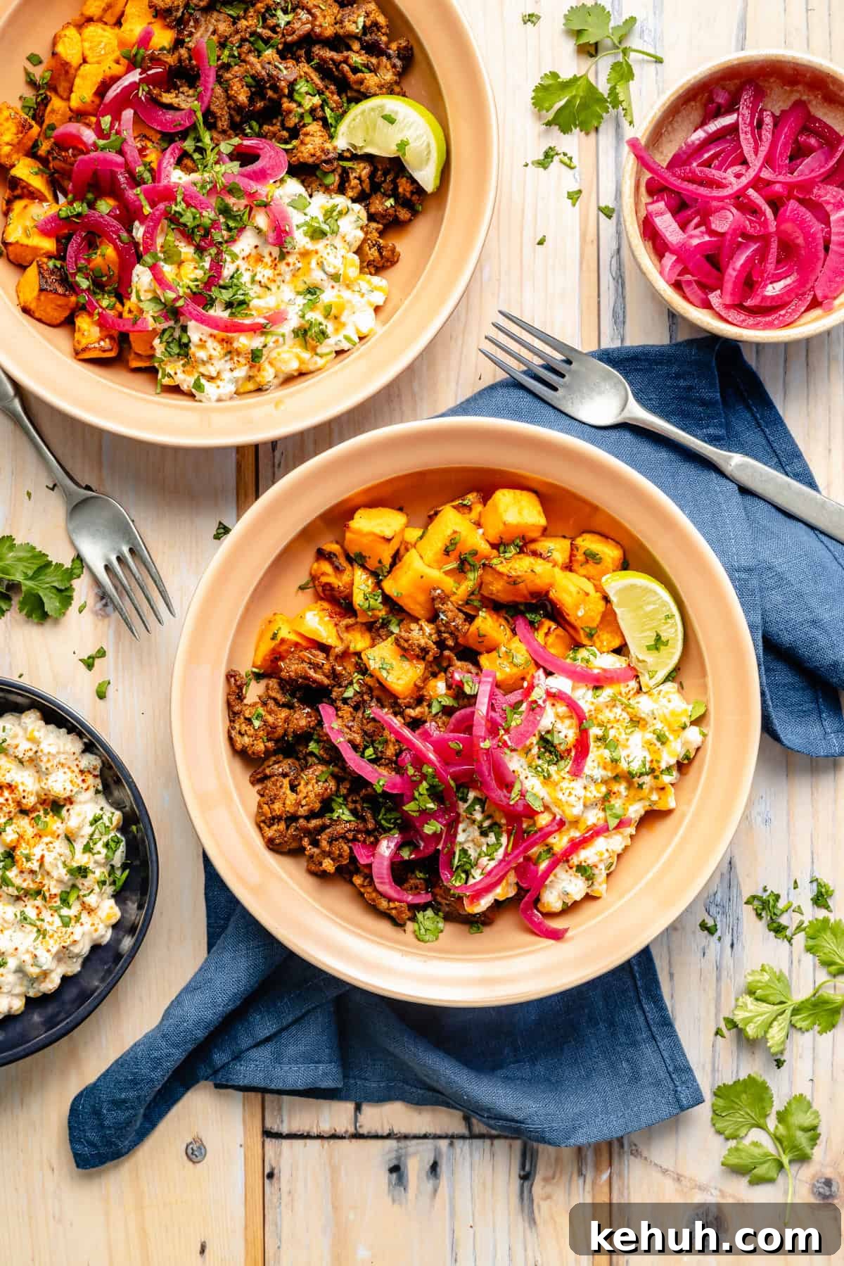 Street Corn & Sweet Potato Beef Bowl Recipe — Flavor-Packed Meal 2 Two bowls filled with ground meat, roasted sweet potatoes, corn, pickled red onions, cilantro, and lime wedges, set on a wood table with a blue napkin, forks, and a small bowl of extra pickled onions nearby.