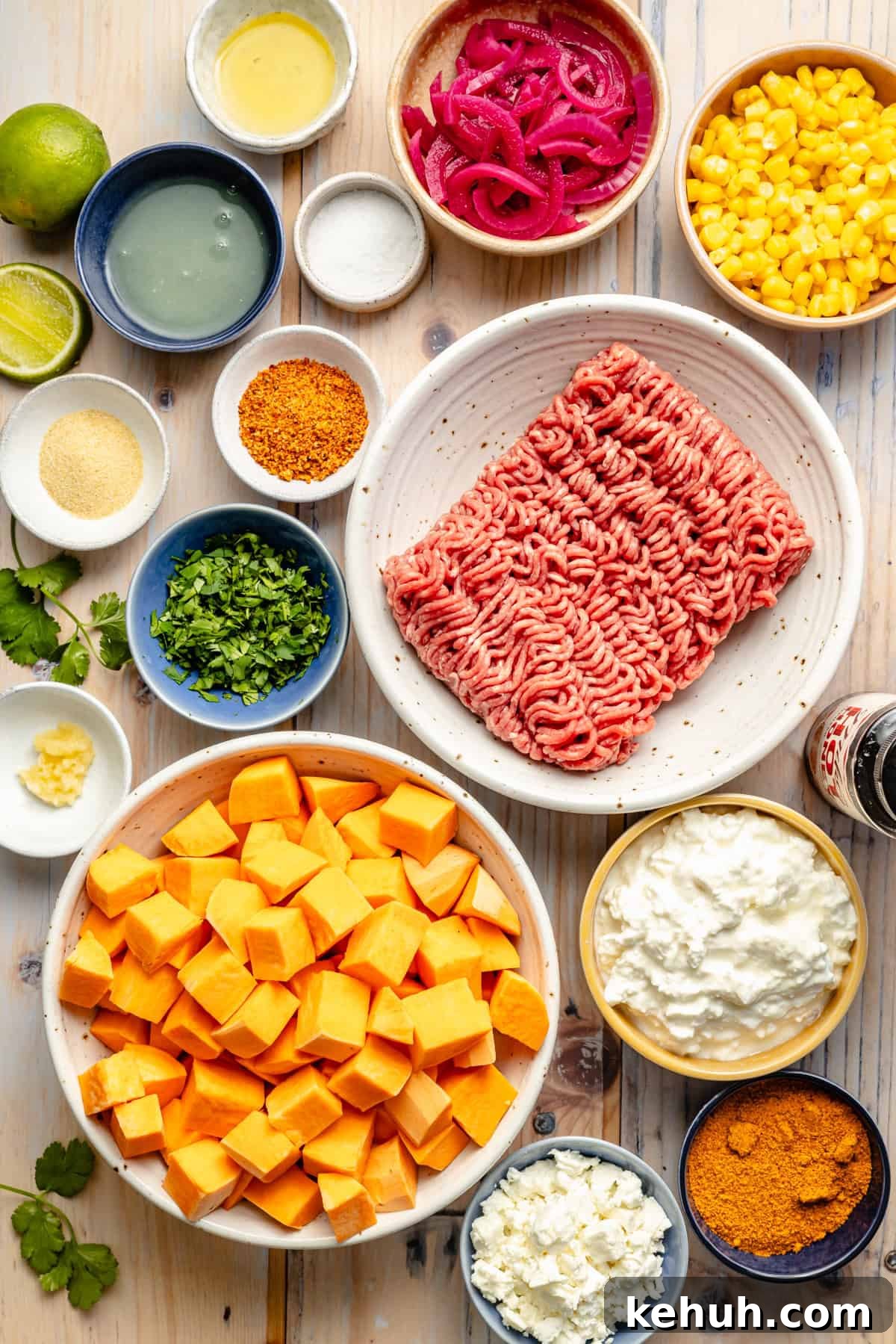 Street Corn & Sweet Potato Beef Bowl Recipe — Flavor-Packed Meal 3 Overhead view of various ingredients in bowls on a wooden surface, including ground beef, cubed sweet potatoes, corn, pickled onions, cottage cheese, lime halves, cilantro, feta, and assorted spices.