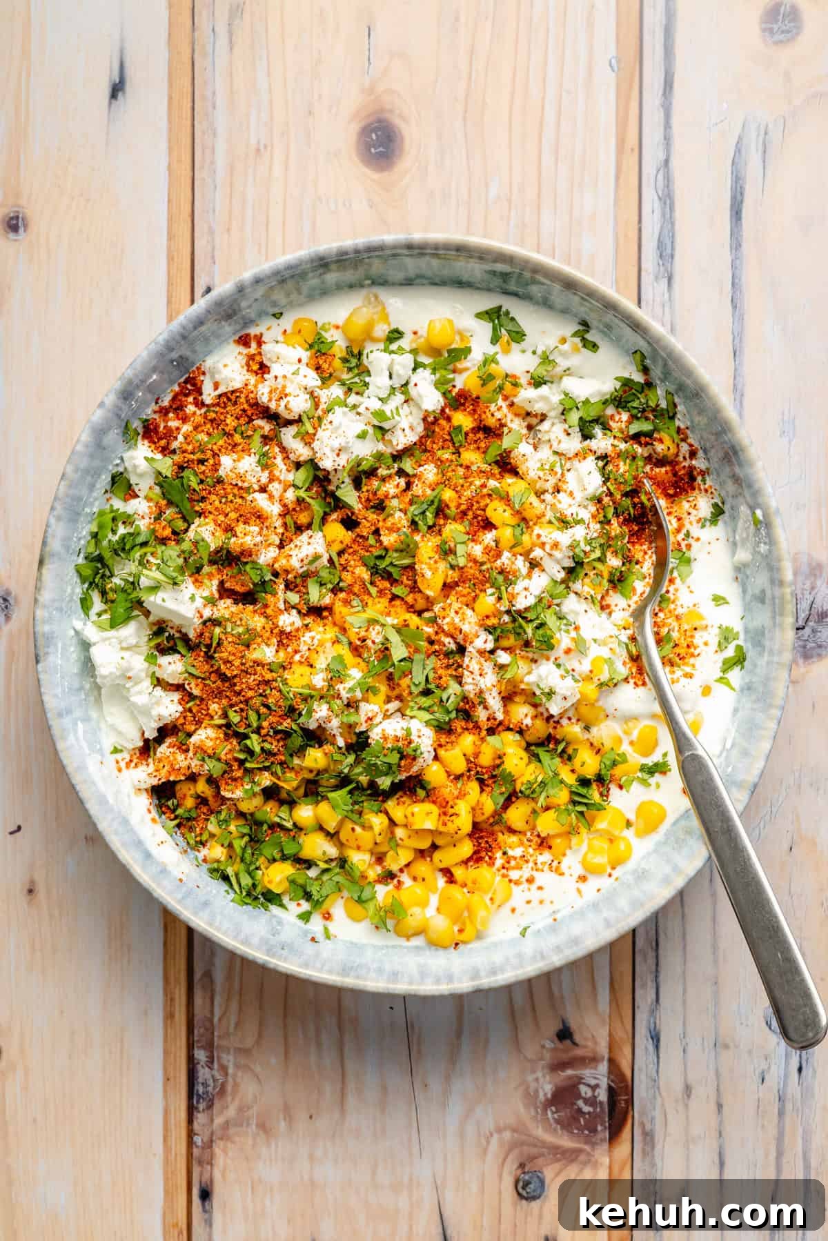 Street Corn & Sweet Potato Beef Bowl Recipe — Flavor-Packed Meal 5 A bowl filled with corn, crumbled cheese, chopped cilantro, and red chili powder on a creamy base, with a spoon placed inside. The bowl sits on a light wooden surface.