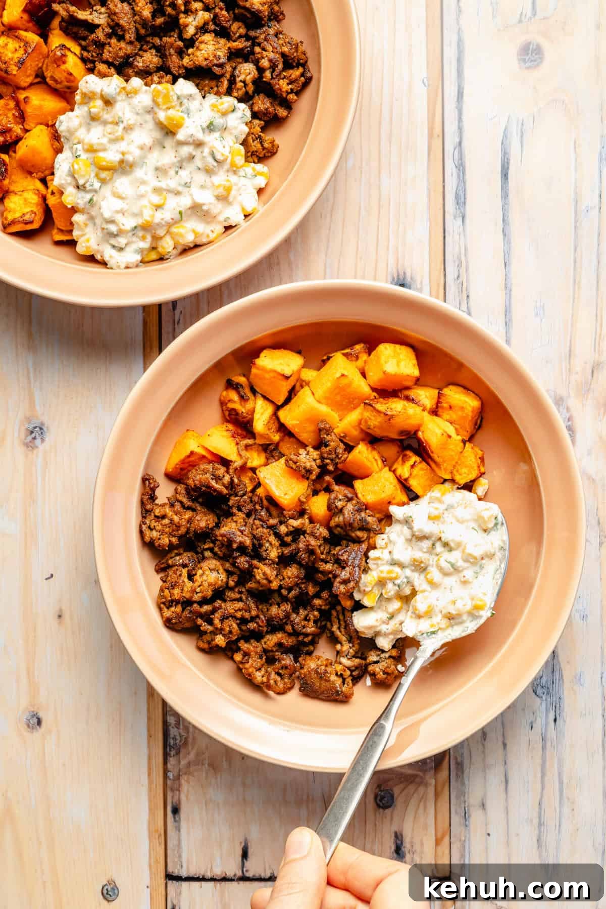 Street Corn & Sweet Potato Beef Bowl Recipe — Flavor-Packed Meal 7 Two beige bowls with roasted sweet potatoes and browned ground meat, topped with a creamy corn mixture. A hand holds a spoon, scooping the corn topping onto one bowl on a light wooden table.