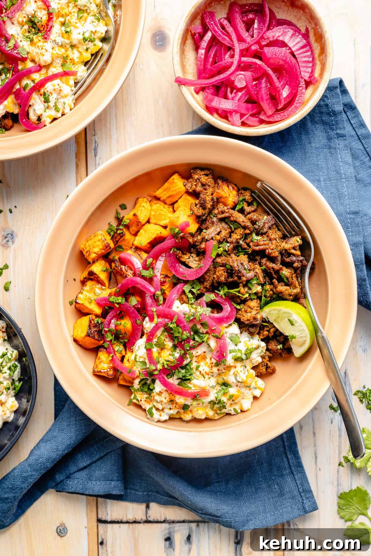 Street Corn & Sweet Potato Beef Bowl Recipe — Flavor-Packed Meal 8 A bowl with seasoned ground meat, roasted sweet potatoes, Mexican street corn salad, and pickled red onions, garnished with cilantro and a lime wedge. A fork rests on the side; other bowls and onions are nearby on a wooden table.