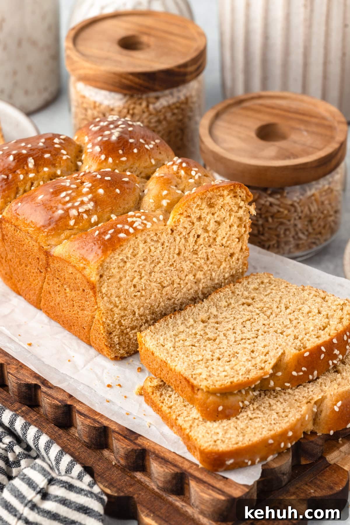 Braided sesame bread loaf on parchment with two slices cut in front. Jars and cloth nearby.