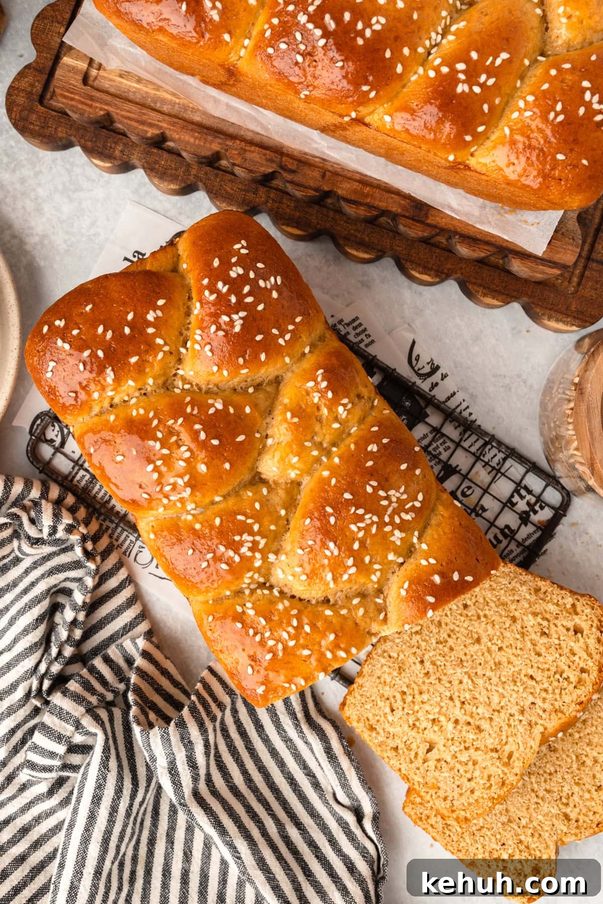 A golden-brown braided sesame loaf with slices sits on a cooling rack near extra bread and cloth.