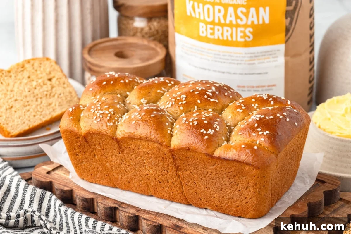 Golden brown sesame-topped bread loaf on parchment and wooden board, sliced bread and butter behind.