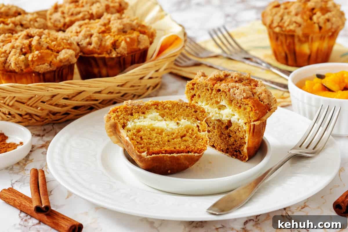 A Pumpkin Cream Cheese Muffins cut in half on a plate reveals a creamy filling, with more muffins in a basket and a bowl of diced fruit in the background.