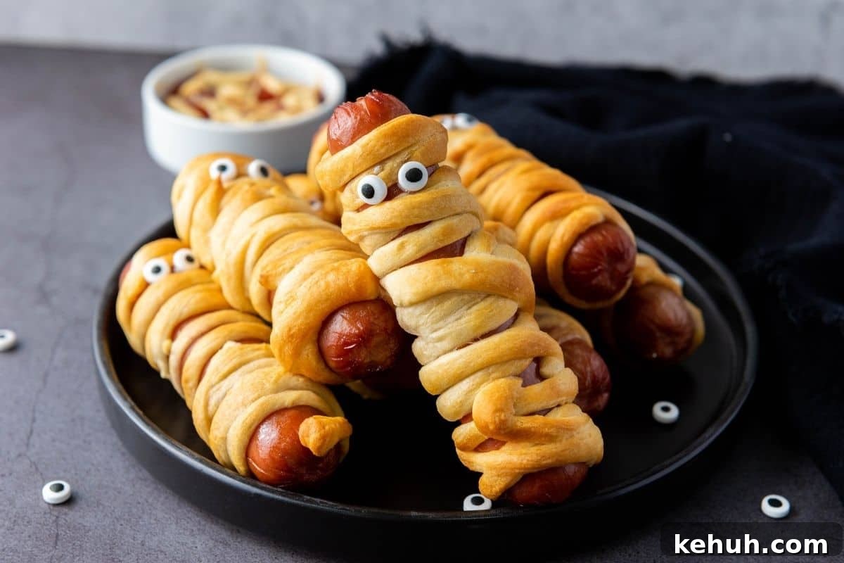 Hot dogs wrapped in crescent dough to resemble mummies, topped with candy eyes, arranged on a black plate with a small bowl of dipping sauce in the background.