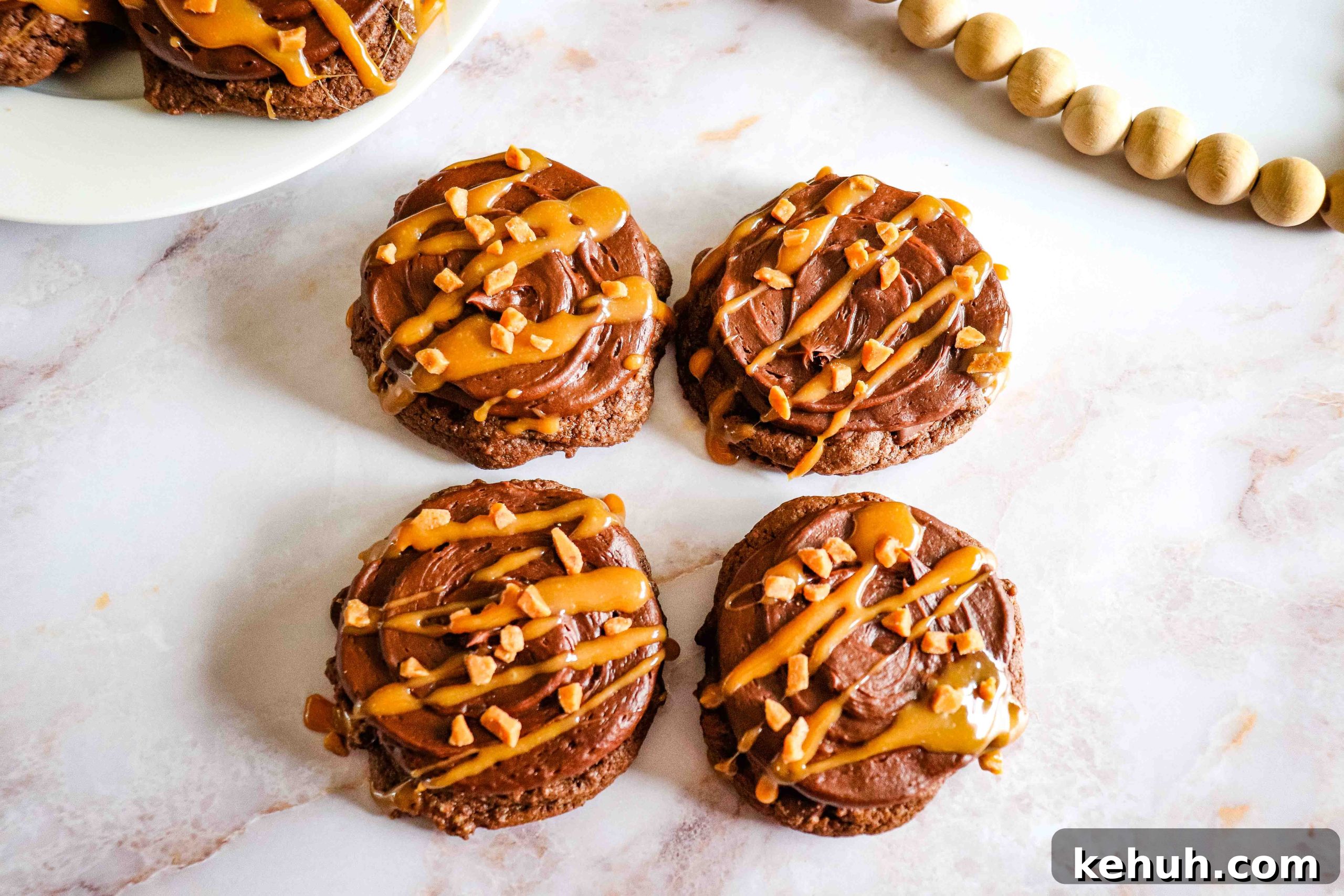 Chocolate Caramel Cookies with frosting, caramel drizzle, and chopped nuts on a white marble surface.