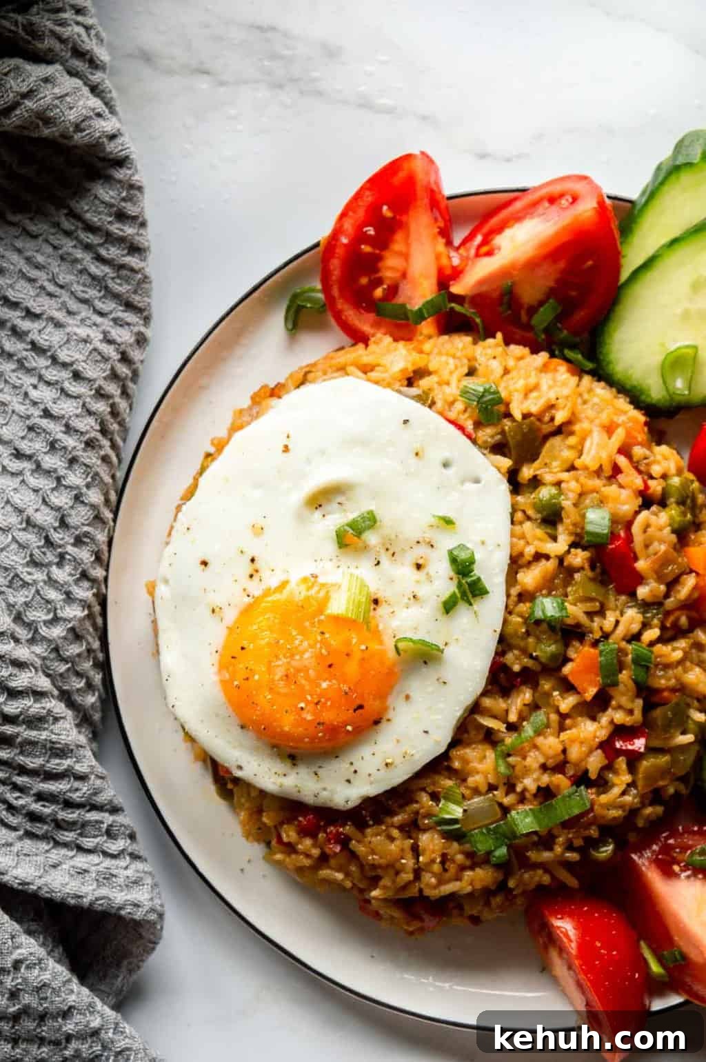 Top down view of a white plate with a round pat of fried rice, a fried egg, and some tomatoes and cucumbers on the edge